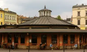 Spokojny rynek na Kazimierzu w Krakowie. Ludzie siedzą przy stolikach przed ceglanym budynkiem z charakterystyczną kopułą.