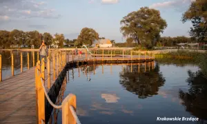 Drewniany pomost nad wodą w największym parku w Krakowie. Ojciec z dzieckiem na ramionach spaceruje po pomoście.