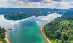 Malownicze jezioro otoczone lasami, idealne miejsce, gdzie na wakacje nad jeziorem. Widać łodzie i małą plażę.