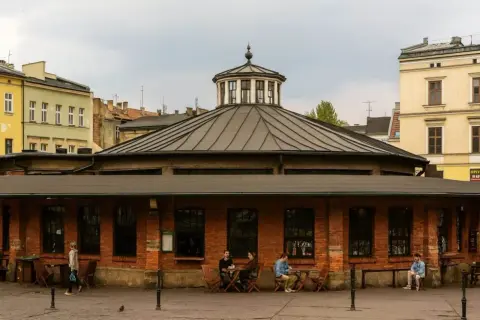 Spokojny rynek na Kazimierzu w Krakowie. Ludzie siedzą przy stolikach przed ceglanym budynkiem z charakterystyczną kopułą.