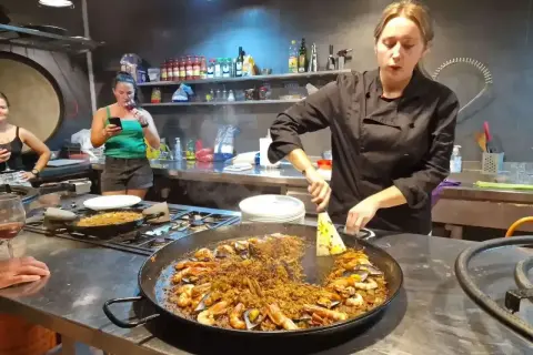 Kobieta w czarnym fartuchu przygotowuje na kuchence ogromną patelnię z owocami morza i ryżem. To musi być najlepsza paella w Barcelonie!