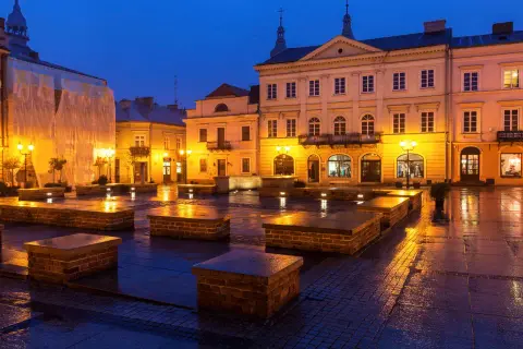 Piotrków Trybunalski atrakcje: Rynek nocą, mokry bruk odbija światła latarni i budynków. Widać kamienne ławki i fasady kamienic.