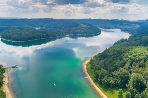 Malownicze jezioro otoczone lasami, idealne miejsce, gdzie na wakacje nad jeziorem. Widać łodzie i małą plażę.