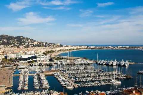 Widok na port w Cannes, gdzie cumują luksusowe jachty. W tle rozciąga się plaża i wzgórza. Idealne miejsce, by zobaczyć Cannes.