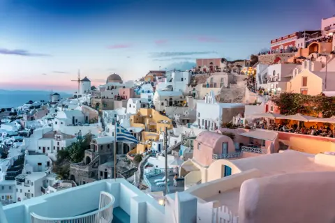 Santorini nocą, z białymi domkami, wiatrakami i grecką flagą, w blasku zachodzącego słońca.