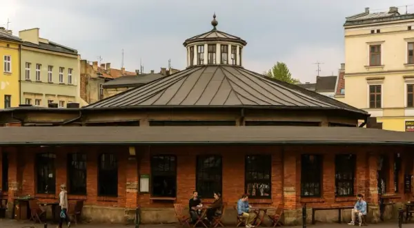Spokojny rynek na Kazimierzu w Krakowie. Ludzie siedzą przy stolikach przed ceglanym budynkiem z charakterystyczną kopułą.