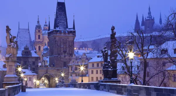 Praga zimą. Śnieg pokrywa Most Karola, wieże i dachy. W oddali widać zarys zamku.