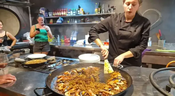 Kobieta w czarnym fartuchu przygotowuje na kuchence ogromną patelnię z owocami morza i ryżem. To musi być najlepsza paella w Barcelonie!