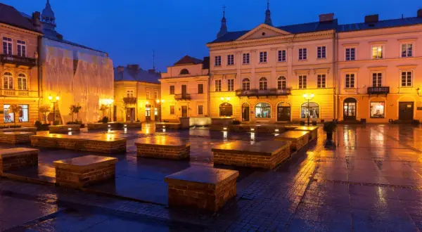 Piotrków Trybunalski atrakcje: Rynek nocą, mokry bruk odbija światła latarni i budynków. Widać kamienne ławki i fasady kamienic.