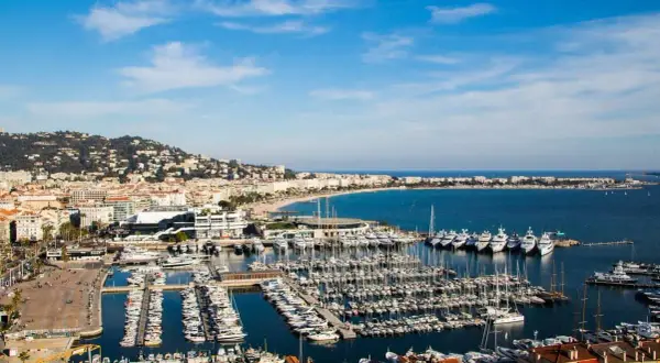 Widok na port w Cannes, gdzie cumują luksusowe jachty. W tle rozciąga się plaża i wzgórza. Idealne miejsce, by zobaczyć Cannes.
