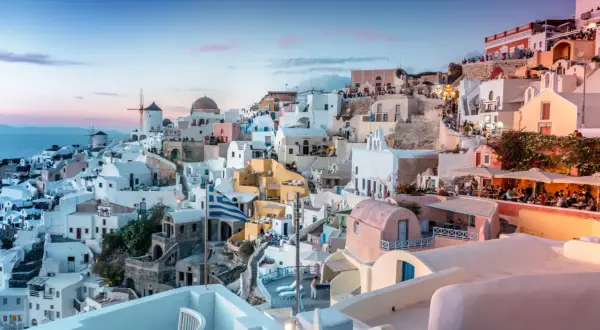 Santorini nocą, z białymi domkami, wiatrakami i grecką flagą, w blasku zachodzącego słońca.