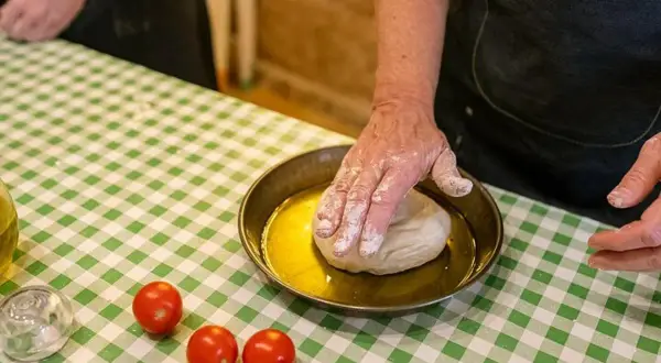 Ręce w mące wyrabiają ciasto na oliwie. Na zielono-białej kratce leżą pomidorki. Pyszne bari jedzenie w przygotowaniu.