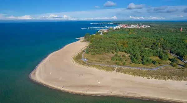 Piękne, piaszczyste plaże i zielone lasy otaczają urokliwe hel miasto. W oddali widać port i zabudowania.