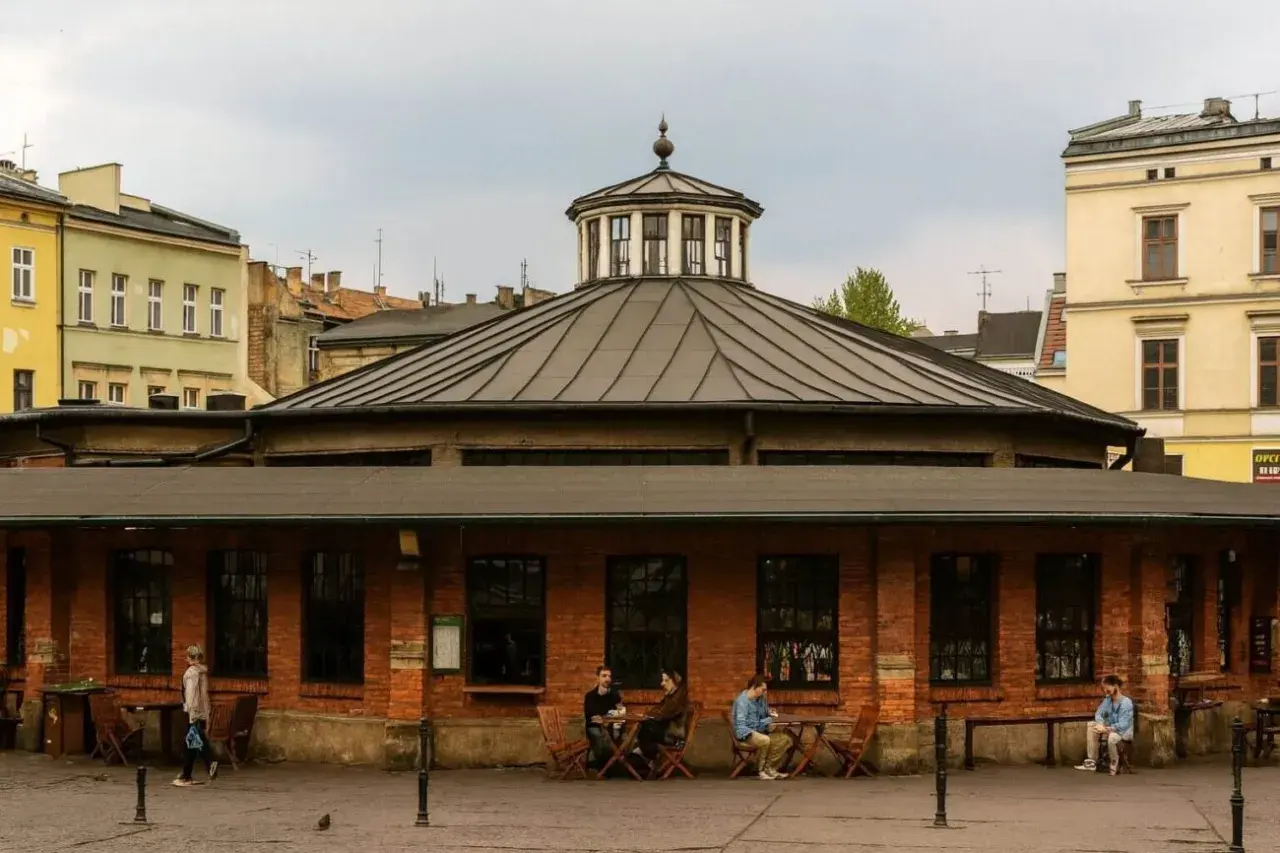 Spokojny rynek na Kazimierzu w Krakowie. Ludzie siedzą przy stolikach przed ceglanym budynkiem z charakterystyczną kopułą.