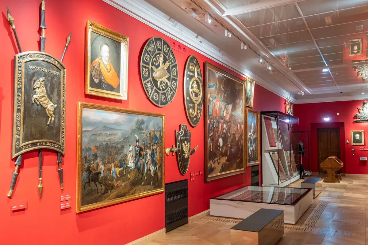 Wnętrze muzeum w Krakowie z obrazami historycznymi, portretem i zbrojami na czerwonych ścianach.