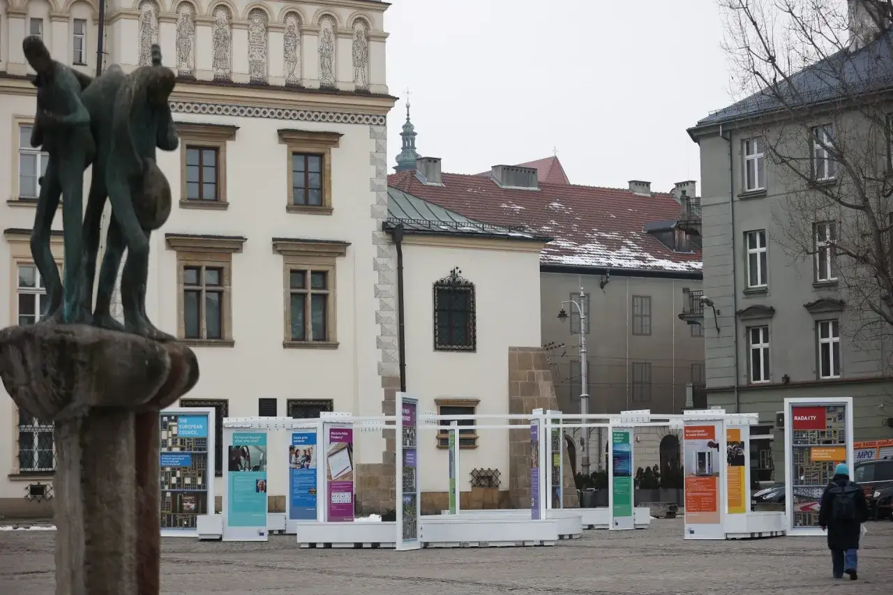Rzeźba na rynku na Kazimierzu w Krakowie, otoczona białymi stoiskami wystawowymi z plakatami.