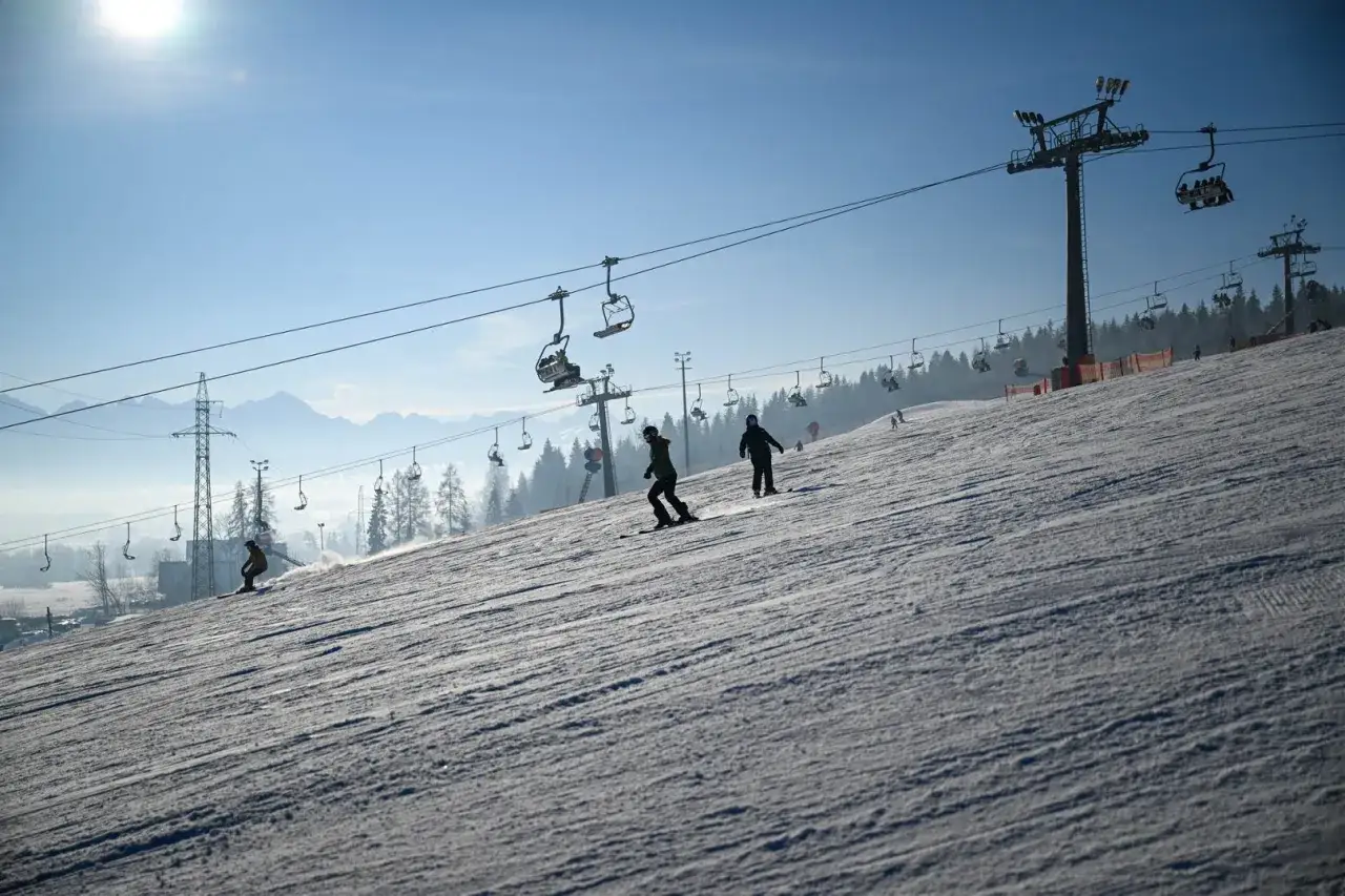 Narciarze i snowboardziści szusują po ośnieżonym stoku, a nad nimi widać wyciągi krzesełkowe. To wspaniałe białka tatrzańska atrakcje zimą.