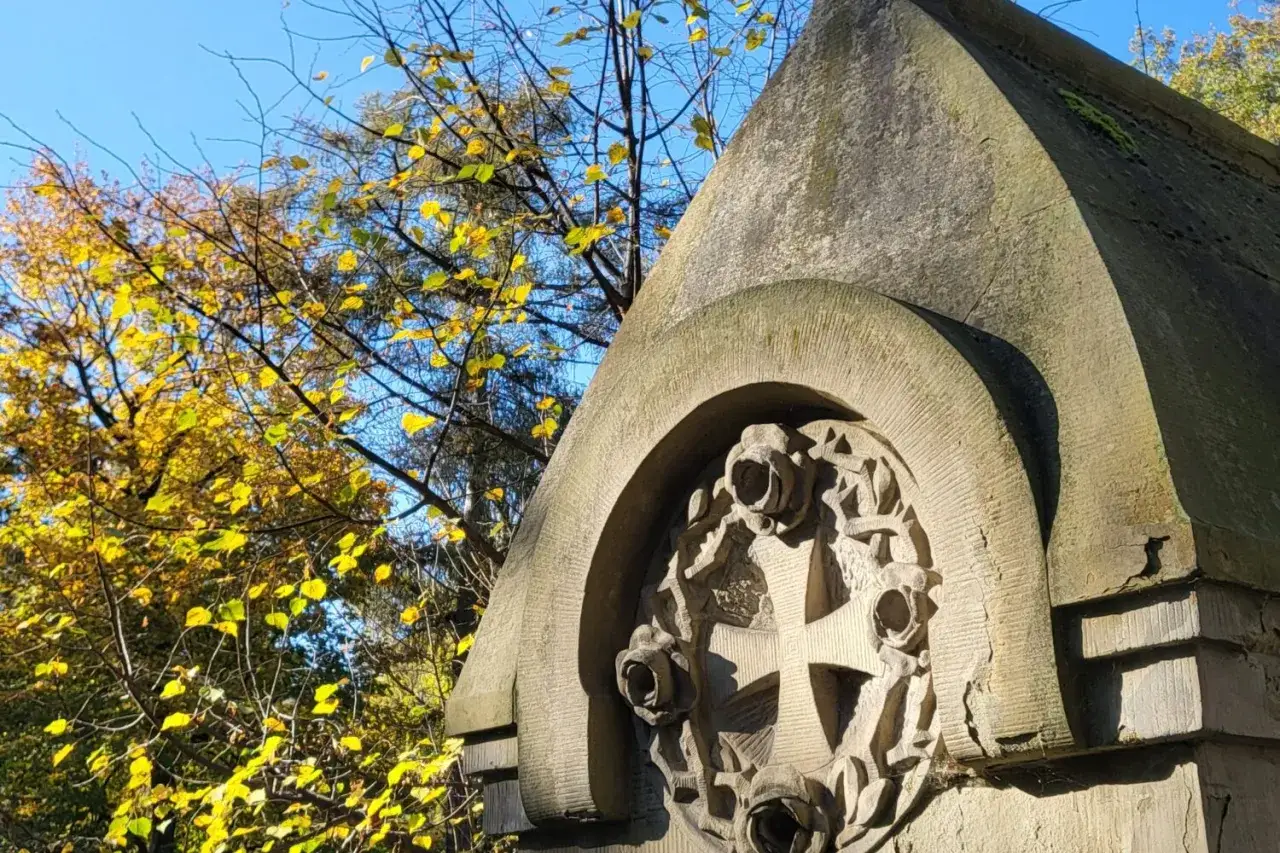 Kamienna rzeźba krzyża z różami na cmentarzu Rakowickim. Jesienne liście drzew tworzą barwne tło.