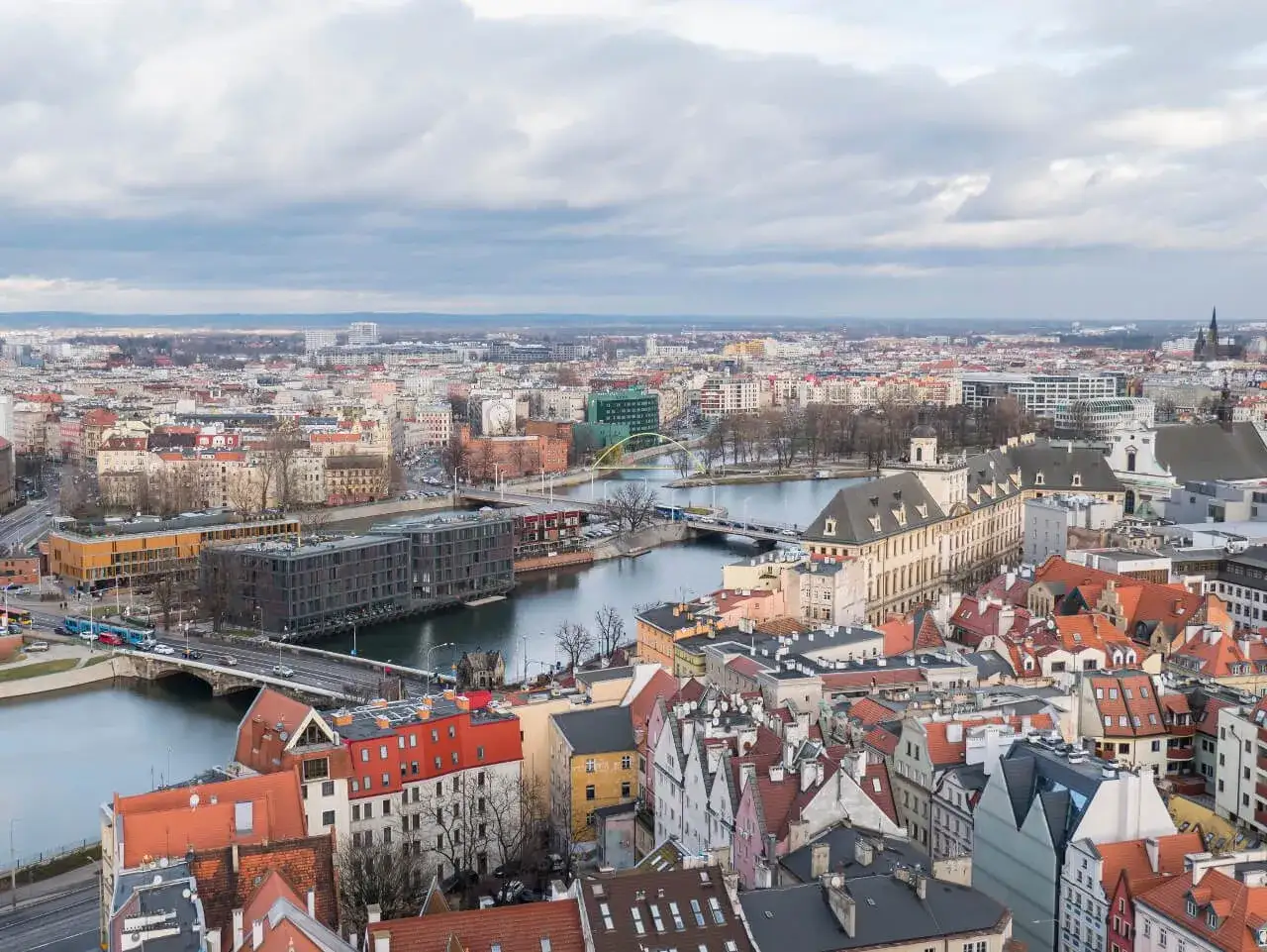 Widok na Wrocław z góry, z rzeką i mostami. Miasto z licznymi dachami i nowoczesnymi budynkami, przypomina widok z jednej z wież widokowych Dolnego Śląska.