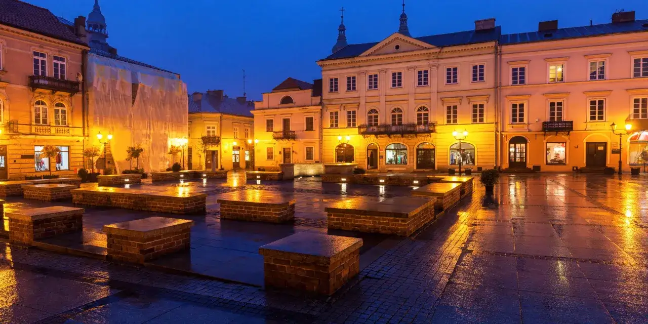 Piotrków Trybunalski atrakcje: Rynek nocą, mokry bruk odbija światła latarni i budynków. Kamienne ławki tworzą ciekawe formy.