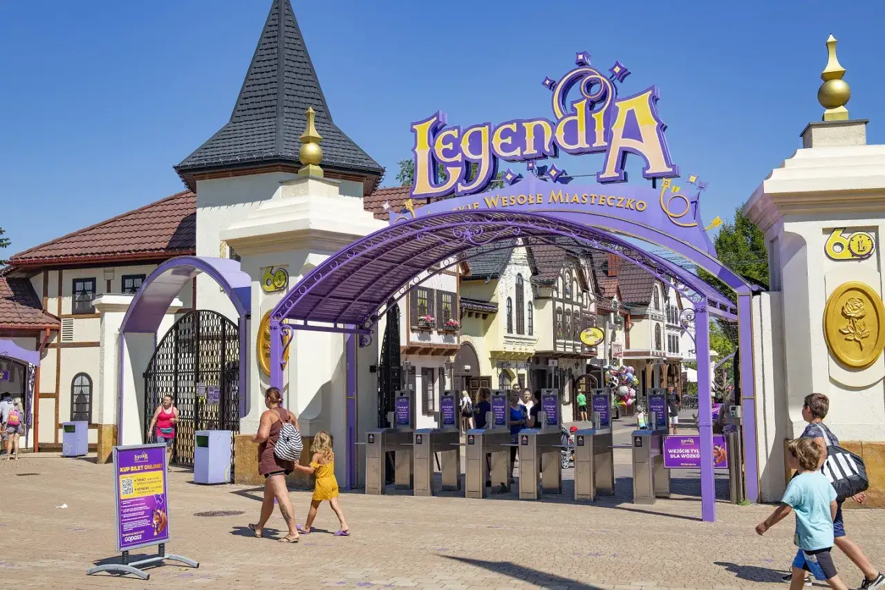 Wejście do Legendii, wesołego miasteczka w Krakowie. Rodziny wchodzą przez bramki pod fioletowym łukiem z napisem 