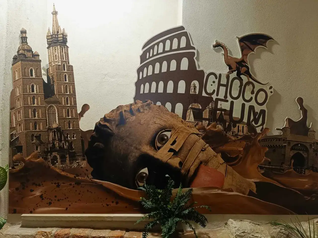 Kraków w czekoladzie! Monumentalna głowa tonie w słodkiej masie, a w tle widać wieże Kościoła Mariackiego i Koloseum. To zapowiedź wizyty w muzeum czekolady Kraków.