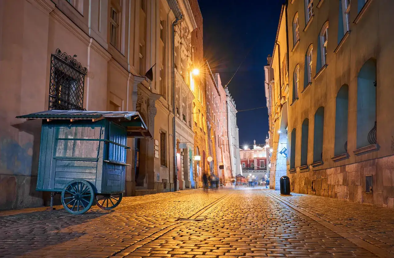 Krak&oacute;w nocą. Brukowana uliczka oświetlona latarniami, z opuszczonym w&oacute;zkiem po lewej i zarysem Sukiennic w oddali.