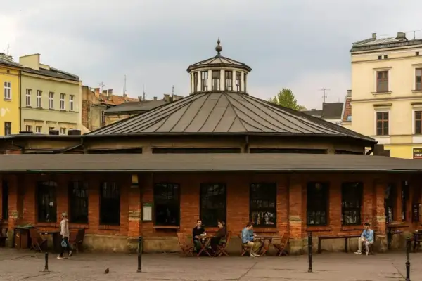 Spokojny rynek na Kazimierzu w Krakowie. Ludzie siedzą przy stolikach przed ceglanym budynkiem z charakterystyczną kopułą.