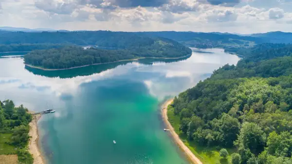 Malownicze jezioro otoczone lasami, idealne miejsce, gdzie na wakacje nad jeziorem. Widać łodzie i małą plażę.